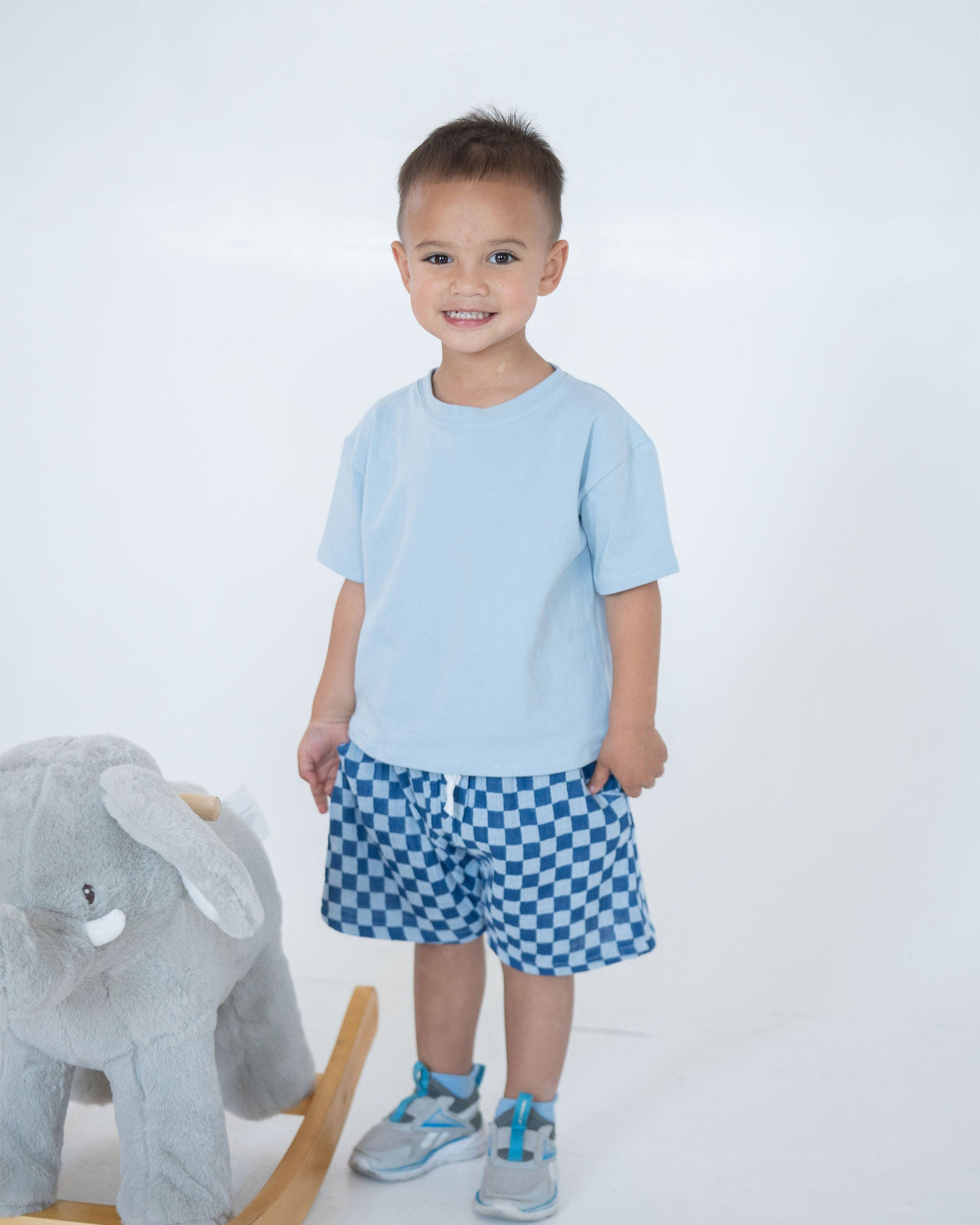 Child wearing a light blue shirt and checkered shorts standing next to a gray elephant toy on a white background