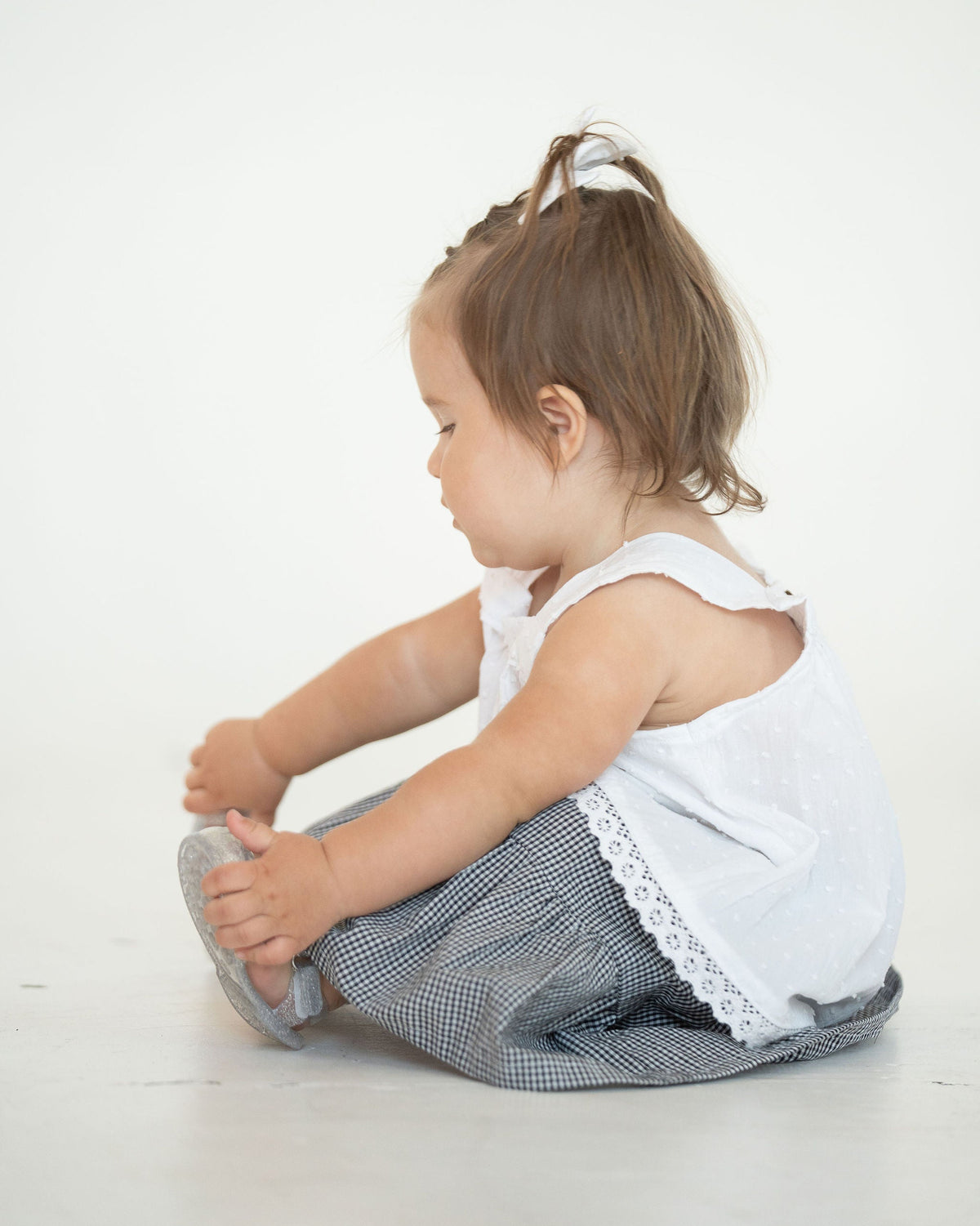 Young child sitting on a white floor wearing a white sleeveless top and checkered skirt