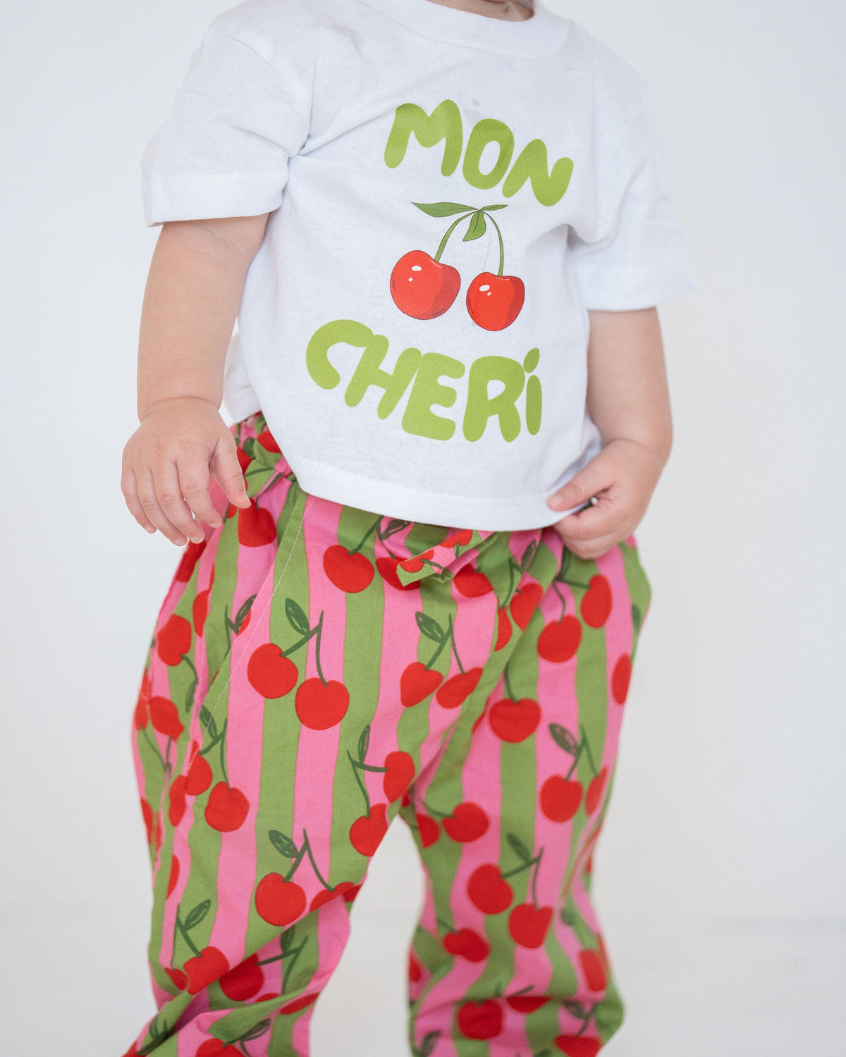 Child wearing a white shirt with 'Mon Cheri' text and colorful cherry-patterned pants on a white background