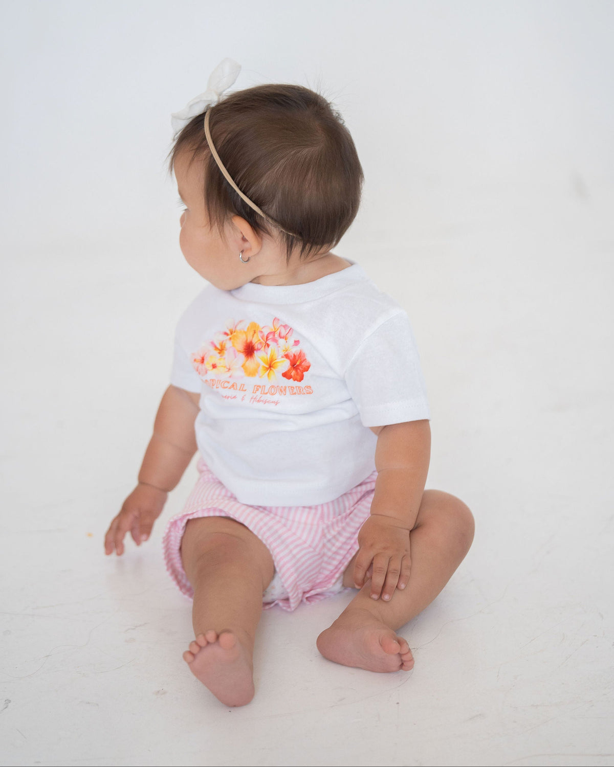 Baby sitting on a white surface wearing a white onesie with a colorful design and text.