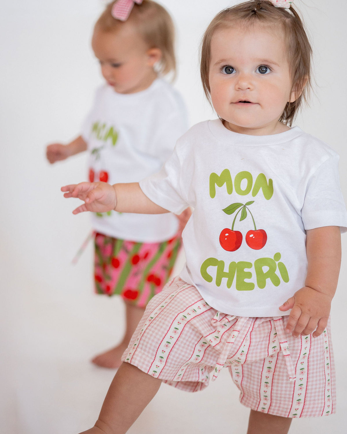 Two young children wearing white t-shirts with colorful text and graphics on a plain background.