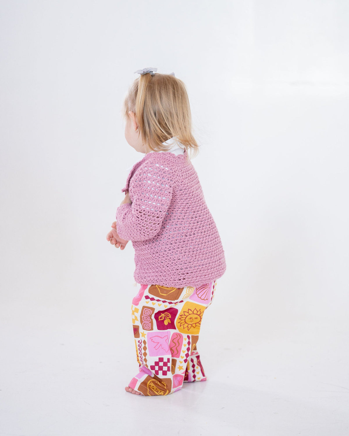 Child wearing a pink crochet cardigan and patterned pants on a white background