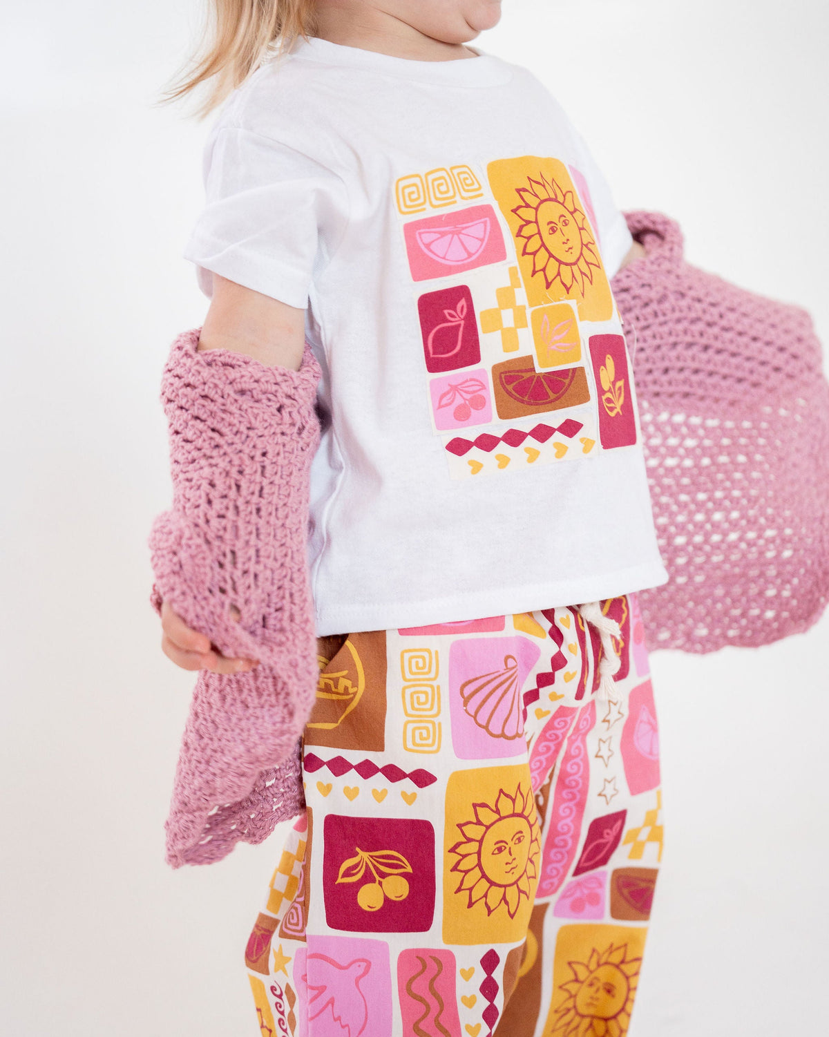 Child wearing a white shirt with colorful graphics and patterned pants, holding a pink knitted bag.