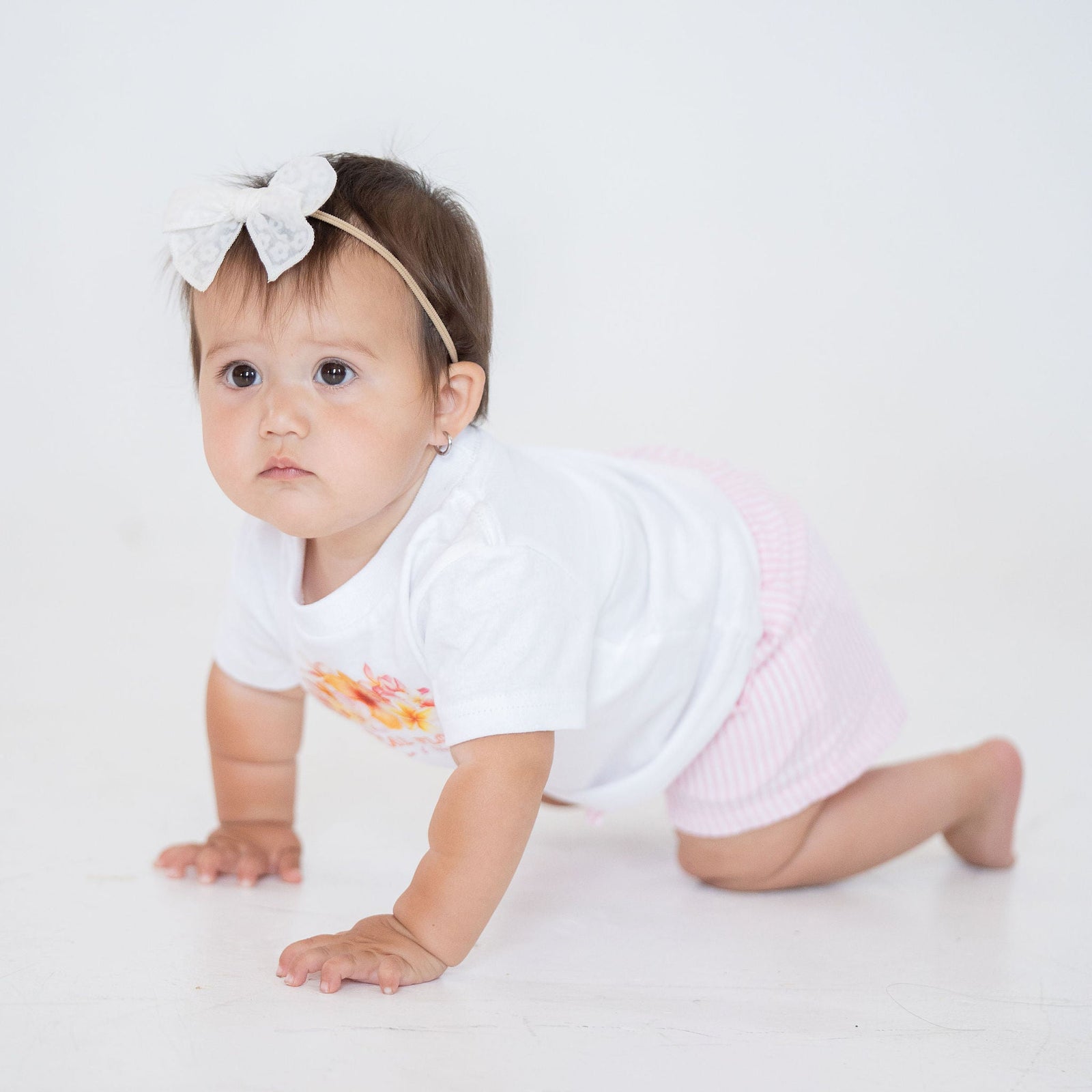 Baby crawling on a white surface wearing a white onesie with colorful design and a headband.